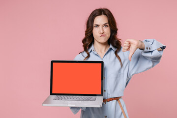 Displeased young brunette woman in casual blue shirt dress hold laptop computer with blank empty screen mock up copy space showing thumb down isolated on pastel pink colour background studio portrait.