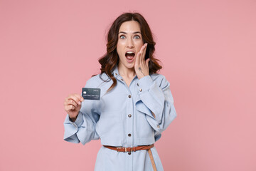 Shocked young brunette woman 20s wearing casual blue shirt dress posing holding in hand credit bank card put hand on cheek looking camera isolated on pastel pink colour background, studio portrait.