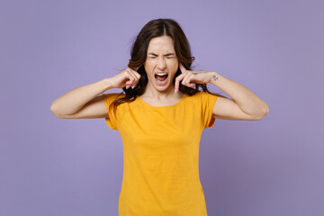 Crazy frustrated young brunette woman 20s wearing basic yellow t-shirt posing covering ears with fingers keeping eyes closed screaming isolated on pastel violet colour background, studio portrait.