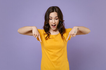 Shocked surprised young brunette woman 20s wearing basic yellow t-shirt posing standing pointing index fingers down on mock up copy space isolated on pastel violet colour background, studio portrait.