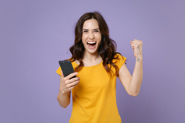 Fototapeta premium Happy young brunette woman 20s wearing basic yellow t-shirt using mobile cell phone typing sms message doing winner gesture looking camera isolated on pastel violet colour background studio portrait.