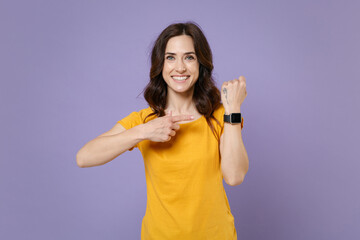 Smiling attractive young brunette woman 20s wearing basic yellow t-shirt posing standing pointing index finger on smart watch on hand isolated on pastel violet colour background, studio portrait.