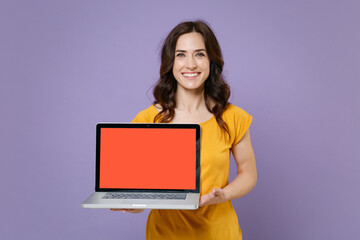 Naklejka premium Smiling young brunette woman 20s in basic yellow t-shirt hold laptop pc computer with blank empty screen mock up copy space looking camera isolated on pastel violet colour background, studio portrait.