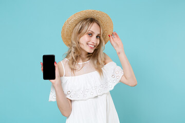Pretty young blonde woman 20s in white summer dress hat hold mobile cell phone with blank empty screen mock up copy space looking camera isolated on blue turquoise colour background studio portrait.