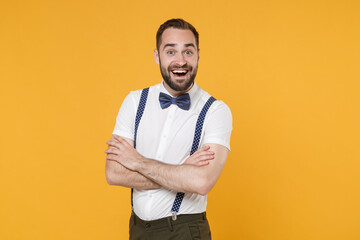 Excited young bearded man 20s wearing white shirt bow-tie suspender posing holding hands crossed keeping mouth open looking camera isolated on bright yellow color wall background, studio portrait.