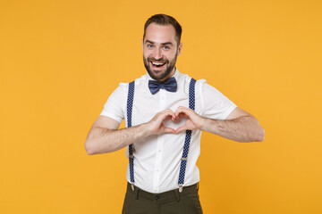 Cheerful funny handsome young bearded man 20s wearing white shirt bow-tie suspender showing shape heart with hands, heart-shape sign looking camera isolated on yellow color background studio portrait.