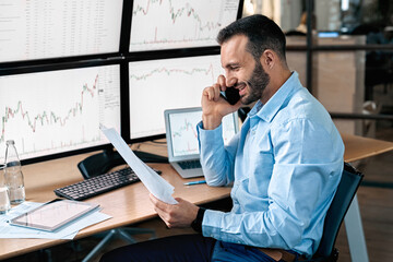 Stock Traiding. Trader sitting in front of monitors with data at office talking on phone doing online trading training reading document smiling cheerful