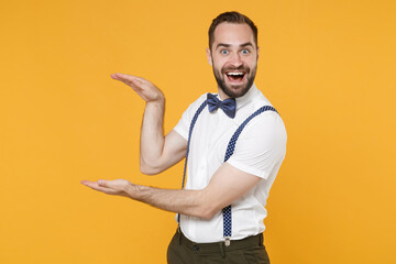 Excited young bearded man 20s in white shirt bow-tie suspender posing standing gesturing demonstrating size with vertical workspace isolated on bright yellow color wall background studio portrait.