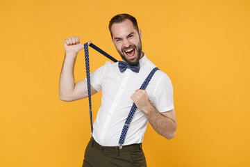 Cheerful funny screaming young bearded man 20s wearing white shirt bow-tie posing standing stretching suspender looking camera isolated on bright yellow color wall background studio portrait.