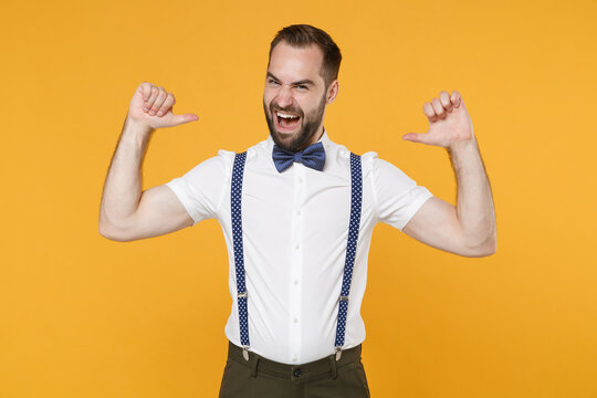 Cheerful funny young bearded man 20s in white shirt bow-tie suspender posing standing pointing thumbs on herself looking camera isolated on bright yellow color wall background studio portrait.