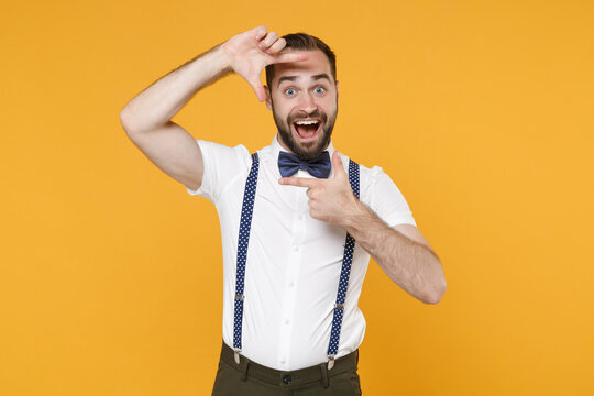 Excited young bearded man 20s in white shirt bow-tie suspender posing standing making hands photo frame gesture keeping mouth open isolated on bright yellow color wall background studio portrait.