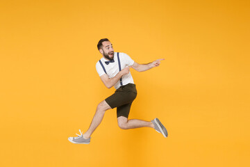 Fototapeta na wymiar Full length side view portrait of excited young bearded man 20s in white shirt suspender shorts posing jumping pointing index fingers aside on mock up copy space isolated on yellow background studio.