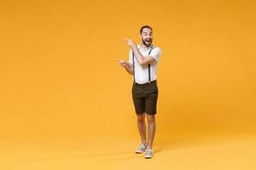 Fototapeta na wymiar Full length portrait of excited young bearded man 20s wearing white shirt suspender shorts posing pointing index fingers aside on mock up copy space isolated on bright yellow color background studio.
