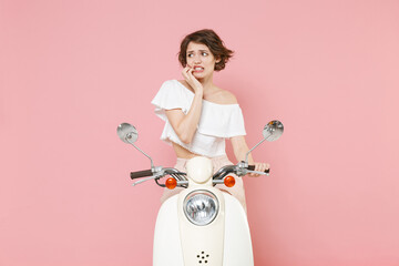 Preoccupied nervous worried young brunette woman 20s wearing white summer clothes gnawing nails looking aside sitting and driving moped isolated on pastel pink colour background studio portrait.