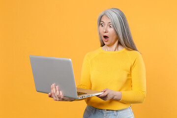 Shocked amazed beautiful gray-haired asian woman in casual clothes standing hold in hands working on laptop pc computer keeping mouth open isolated on bright yellow colour background, studio portrait.