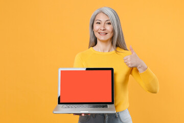 Smiling gray-haired asian woman wearing casual clothes hold laptop pc computer with blank empty screen mock up copy space showing thumb up isolated on bright yellow colour background, studio portrait.