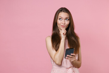 Pensive young brunette woman 20s wearing pink summer dotted dress posing using mobile cell phone typing sms message put hand prop up on chin isolated on pastel pink color background studio portrait.