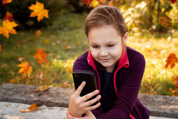 A beautiful girl of eight years old holds a phone in her hands against the background of early autumn.