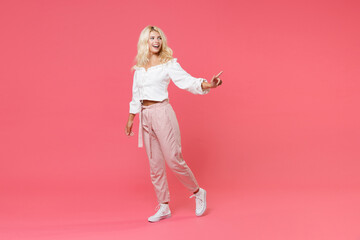Fototapeta na wymiar Full length portrait of cheerful young blonde woman 20s wearing white casual clothes standing pointing index finger aside on mock up copy space isolated on bright pink colour background in studio.