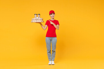 Full length body delivery employee woman in red cap blank t-shirt uniform work courier in service hold bring food order pizza in cardboard flatbox, paper cups of coffee isolated on yellow background.