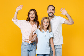 Obraz premium Laughing young parents mom dad with child kid daughter teen girl in basic t-shirts waving and greeting with hand as notices someone isolated on yellow background studio portrait. Family day concept.