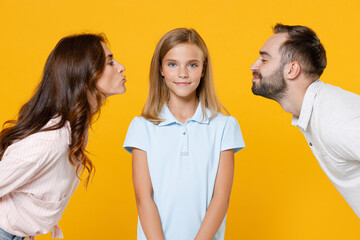 Pretty smiling young happy parents mom dad with child kid daughter teen girl in basic t-shirts kissing in cheeks isolated on yellow background studio portrait. Family day parenthood childhood concept.