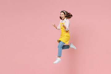 Full length portrait of excited young woman housewife 20s in yellow apron jumping pointing index fingers aside doing housework isolated on pastel pink colour background studio. Housekeeping concept.