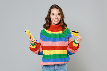 Smiling beautiful young brunette woman 20s in casual colorful knitted sweater standing using mobile cell phone hold credit bank card looking camera isolated on grey colour background, studio portrait.