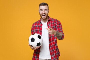 Funny excited young man football fan in basic shirt cheer up support favorite team with soccer ball pointing index finger on camera isolated on yellow background studio. People sport leisure concept.