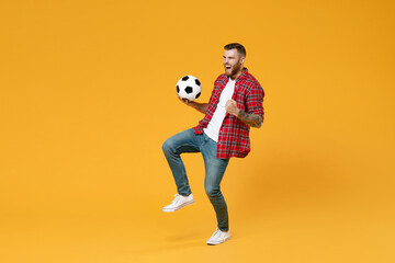 Full length portrait happy young man football fan in red shirt cheer up support favorite team with soccer ball doing winner gesture isolated on yellow background studio. People sport leisure concept.