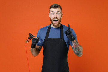Fototapeta na wymiar Excited professional tattooer master artist man in blue t-shirt apron hold machine black ink in jar, equipment for making tattoo art on body point finger up with new idea isolated on brown background.