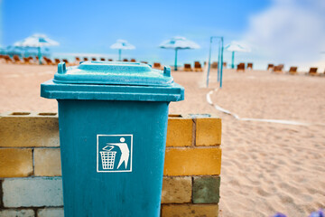 Trash can on the beach. Plastic large waste tank. Maintaining cleanliness in the recreation area of people. Trash sign. Fence made of brick blocks. Beach with umbrellas and sun loungers.