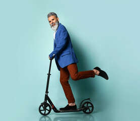 Gray-haired elderly man in white shirt, jacket, brown pants, loafers. Riding black scooter and posing sideways on blue background