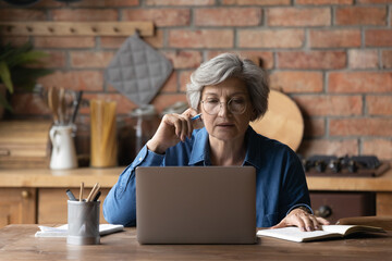 Smart senior grey-haired woman in glasses sit at table at home kitchen look at laptop screen...