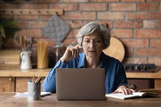 Smart senior grey-haired woman in glasses sit at table at home kitchen look at laptop screen working online. Mature Caucasian 60s female study or watch webinar on computer using internet connection.