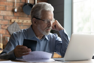 Stressed elderly grey-haired man in glasses look at laptop screen have problems paying bills taxes online. Thoughtful mature 70s male manage household finances, calculate expenses expenditures at home