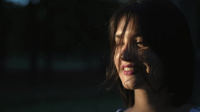 Cheery girl with long forelock standing and smiling in a park in summer in slo-mo 