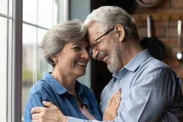 Smiling elderly man and woman hug cuddle touch foreheads show love and care. Happy mature grey-haired Caucasian 60s couple husband and wife embrace, enjoy pleasant family weekend time together.