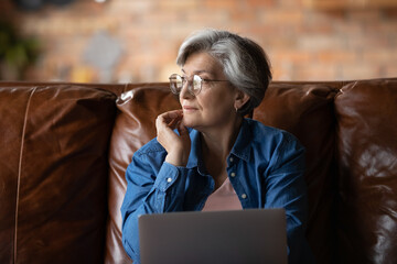 Dreamy elderly Caucasian grey-haired woman in glasses distracted from computer work look in window...