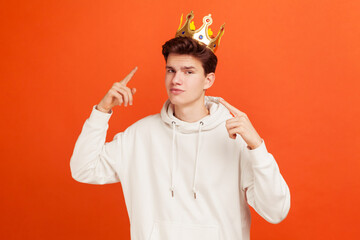 Selfish egoistic teenager in casual style white hoodie pointing fingers on golden crown on his...