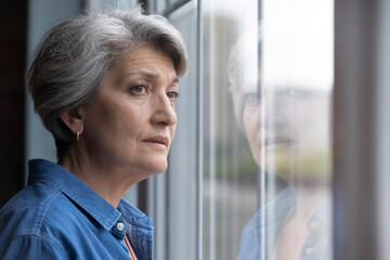Unhappy elderly Caucasian 60s woman look in window distance mourning or yearning at home. Sad mature grey-haired female lost in thoughts feel lonely abandoned in retirement house. Solitude concept.