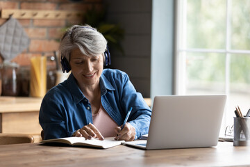 Modern elderly grey-haired 60s woman in headphones take online course or training on computer at...