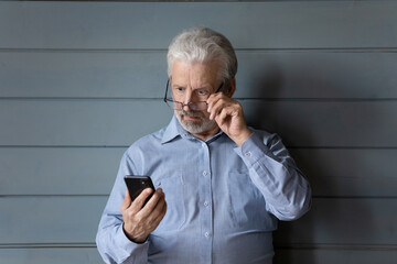 Stunned elderly grey-haired man isolated on grey wooden wall background look at cellphone screen...