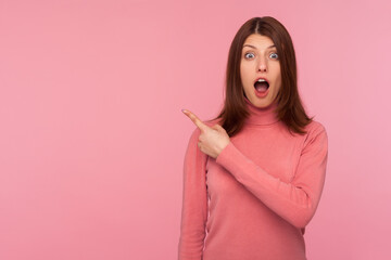 Obraz premium Extremely shocked brunette woman in pink sweater pointing away looking at camera with big eyes and open mouth, surprised with advertising, empty space. Indoor studio shot isolated on pink background