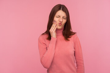 Unhealthy brunette woman touching cheek feeling unpleasant dental pain, oral cavities inflammation, gingivitis. Indoor studio shot isolated on pink background