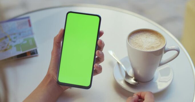 Crop view of female person holding smartphone and pressing on mockup screen while sitting at table with coffee and map on it. Concept of greenscreen and chroma key.