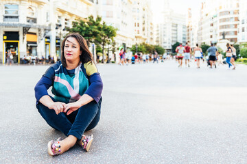 brunette woman in casual clothes sitting on the ground in a square in Europe with people, looking into the distance