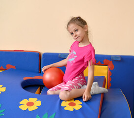 A little girl sits with a ball on a gymnastic mat