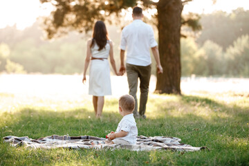 little son sitting on blanket outdoors in the park, mom and dad on background. The concept of summer holiday. Mother's, father's, baby's day. Family spending time together on nature.