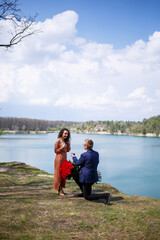 Fototapeta premium Lake in the forest: a man made a surprise to his girlfriend, he puts a ring on her finger, a declaration of love and an offer of marriage. Girl with a smile on her face and a bouquet of red roses